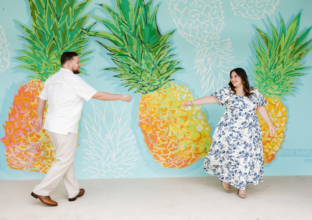 A playful moment as Claire and Reece reach for each other in front of the bright pineapple mural, a fun scene from their engagement shoot in Key Largo.