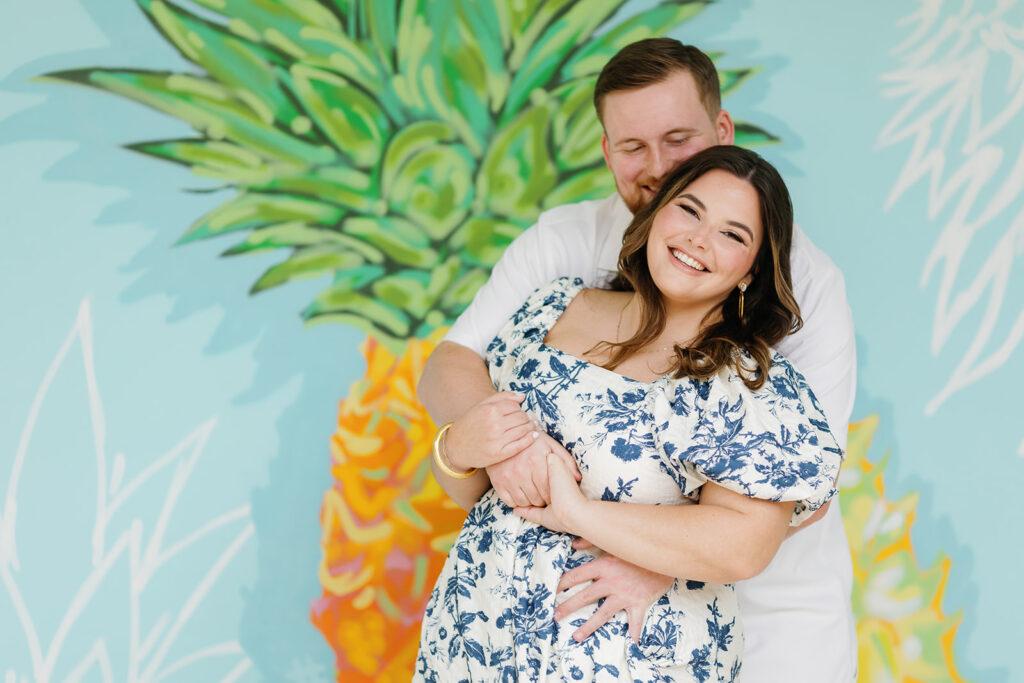 Claire laughs as Reece wraps his arms around her in front of a vibrant pineapple mural at Bakers Cay Resort in Key Largo, Florida. Their engagement session captures the pure joy and connection between them.