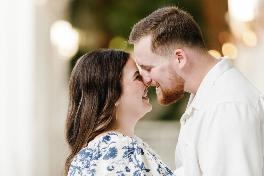 Claire and Reece lean in, forehead to forehead, smiling softly at each other during their engagement session. The warm glow of the lights creates a romantic atmosphere.