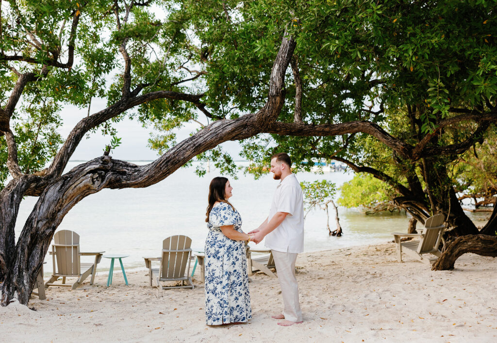 Claire and Reece stand barefoot on the sandy shore at Bakers Cay Resort, exchanging a loving gaze beneath the trees—perfect for beach wedding photography.