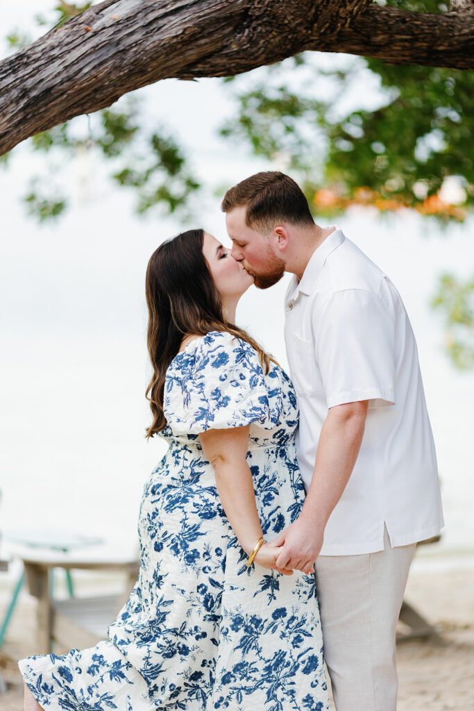 Claire and Reece share a romantic kiss under a tree during their beach engagement session, captured by Claudia Amalia Photography.