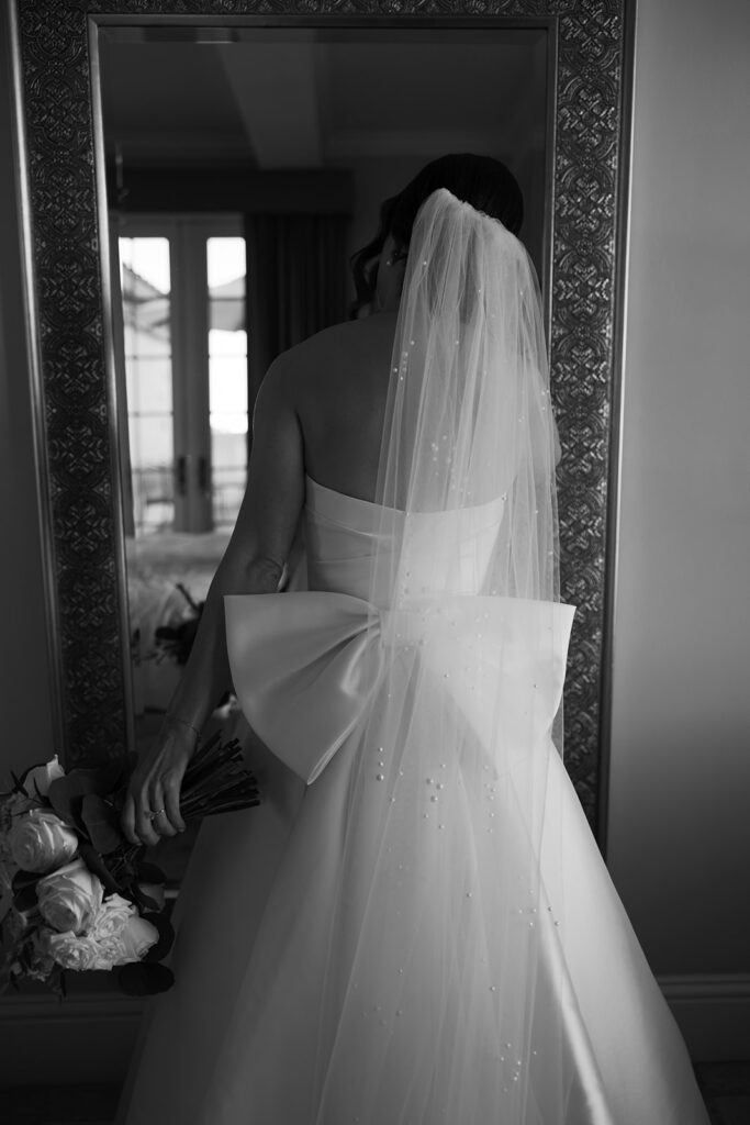 A black-and-white artistic portrait of a bride seen from behind, showcasing her flowing veil, statement bow, and delicate beaded details.