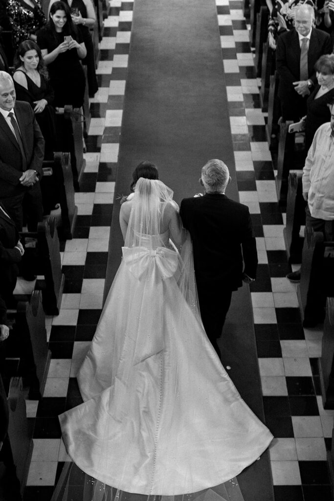 A black-and-white shot of a bride walking down the aisle with her father, taken from behind for a dramatic and elegant perspective.