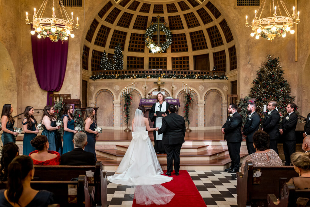 A bride and groom stand before the officiant, exchanging vows in a warmly lit, cathedral-style church with elegant chandeliers.