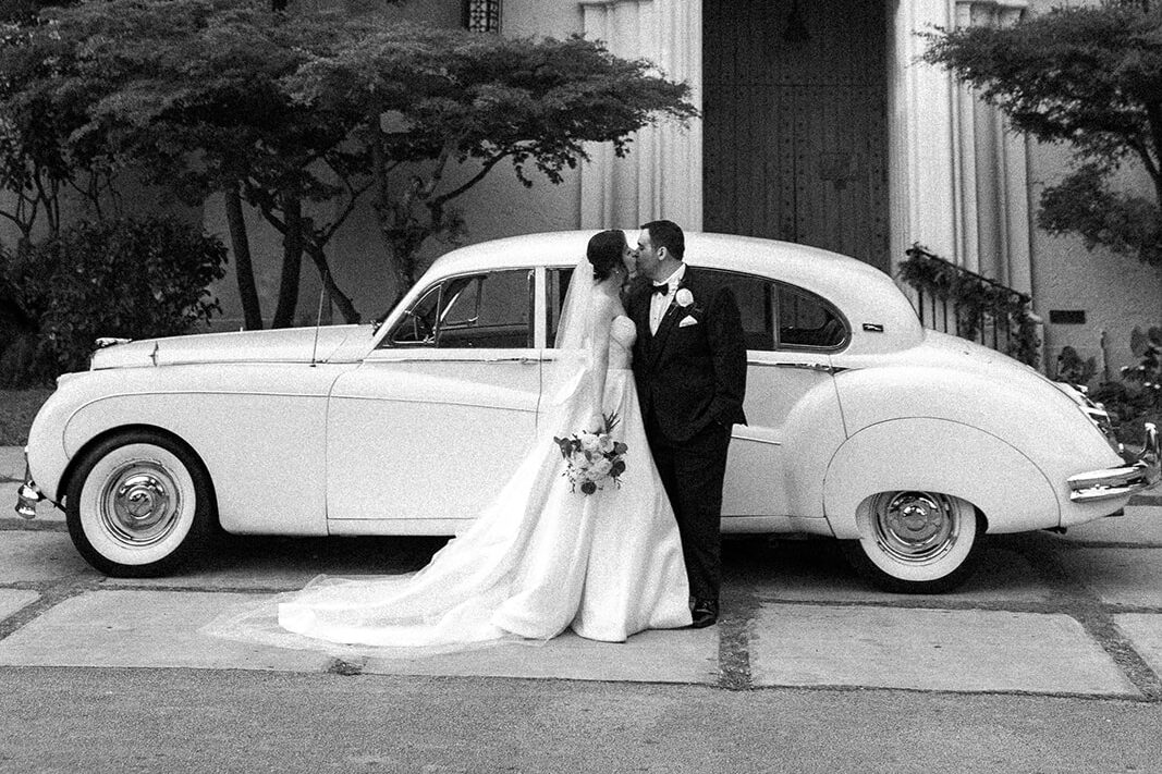 Black and white photo of a bride and groom kissing in front of a vintage car.