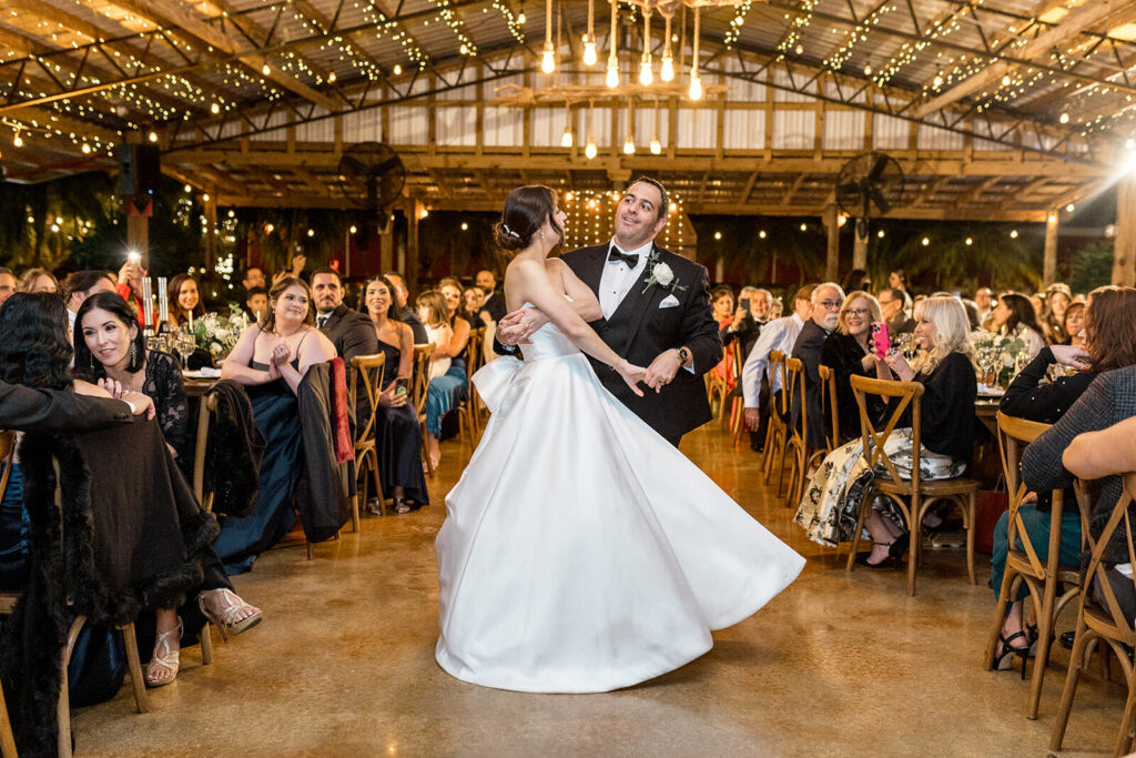 Bride and groom sharing a joyful first dance at their wedding reception, surrounded by family and friends under twinkling string lights.