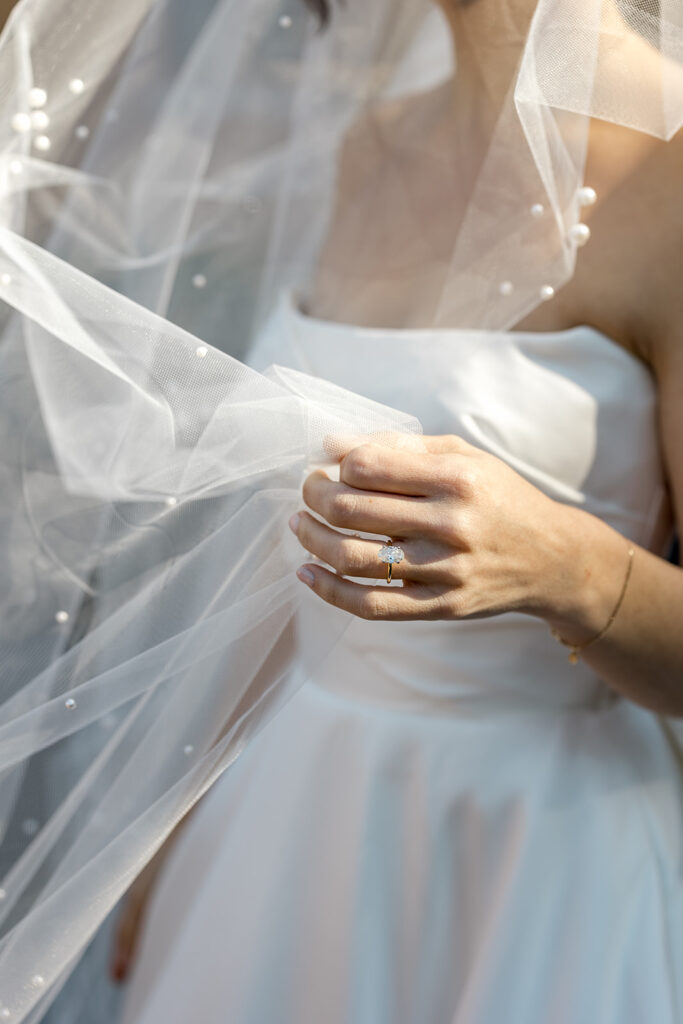 The bride’s delicate hand holding her veil, highlighting her engagement ring and pearl-adorned sheer fabric.