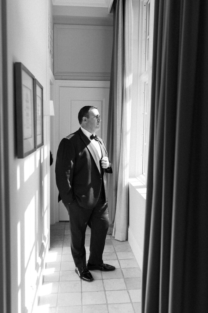 The groom stands by the window, gazing out as sunlight filters through, captured at the Biltmore Hotel.