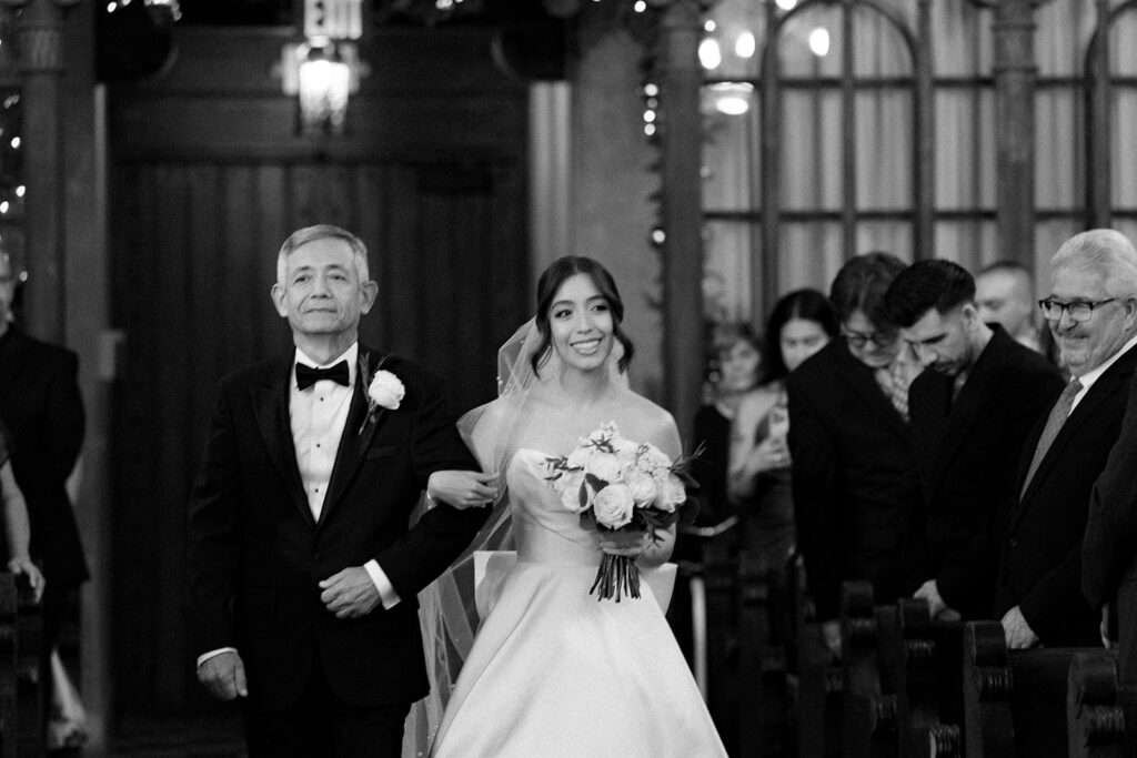 Bride walking down the aisle with her father, smiling as guests watch.