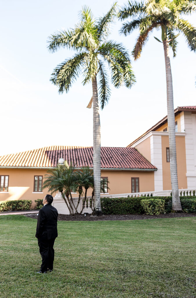 Groom in a black tuxedo stands on a lush lawn, looking up at the palm trees.