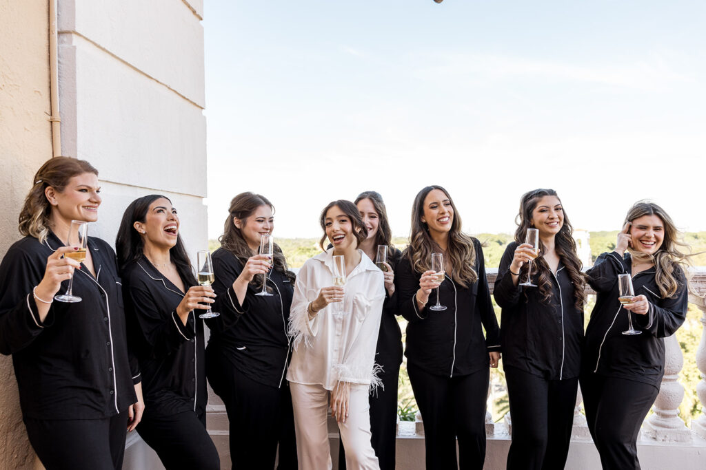 Bride laughing with her bridesmaids in matching black pajamas while holding champagne glasses.